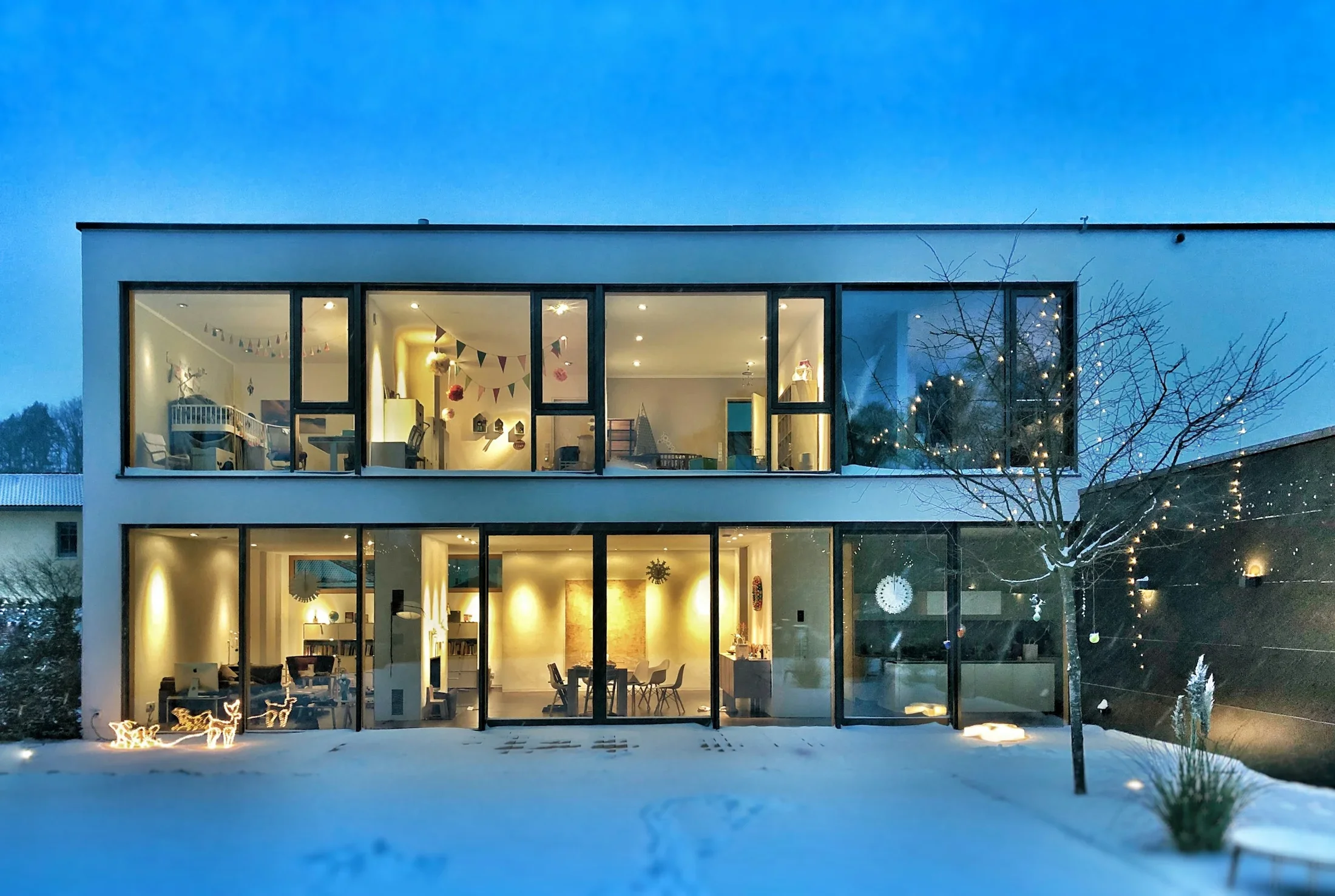 A modern two-story glass-fronted home with warm interior lighting and a snowy garden